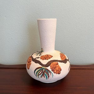 May include: A white ceramic vase with a round base and a narrow neck. The vase is decorated with a brown branch design and clusters of orange-brown pinecones. Green and blue leaves accent the design. The vase sits on a dark wood surface.