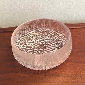May include: A clear, round glass bowl with a textured, ice-like exterior and a bubbly interior pattern. The bowl is set on a dark wooden surface. The bowl's design suggests it could be used for decorative purposes or serving.