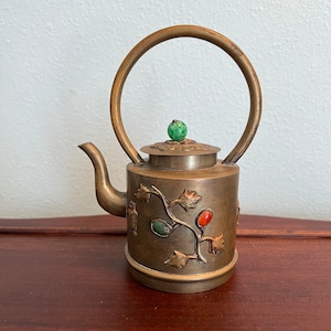 May include: A small, brass teapot with a green jade knob on the lid. The teapot is decorated with a vine design and has several green and red gemstones embedded in the metal.