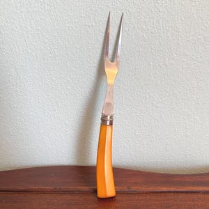 May include: A vintage carving fork with a stainless steel head and a bright orange handle. The handle has a curved shape and a decorative silver band near the head. The fork is resting on a dark wooden surface.