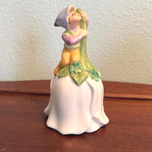 May include: A ceramic figurine of a small child wearing a blue cap and holding onto a green stem. The child is perched atop a white flower-shaped base with green leaves. The figurine has a whimsical, fairytale aesthetic.
