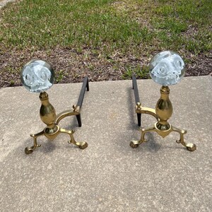 May include: Two antique brass and glass fireplace andirons. Each andiron features a clear glass orb with a white floral design, mounted on a brass stand with a decorative base and a black metal support.