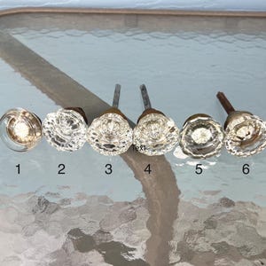 May include: Six clear glass cabinet knobs with varying decorative designs. Each knob has a metal stem. The knobs are numbered 1 through 6. The knobs are displayed on a glass table.