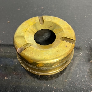 May include: A brass ashtray with four grooves for cigarettes. The ashtray has a circular shape and a smooth, polished surface.