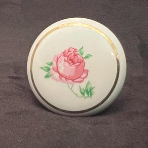 May include: A white ceramic cabinet knob with a pink rose and green leaves design. The knob has a gold-colored trim around the edge of the floral design. The knob is round and has a smooth surface.