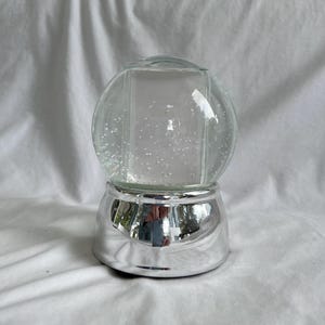 May include: A clear glass snow globe filled with liquid and small white particles. The globe is mounted on a smooth, reflective silver-colored base with a curved, tapered shape. The surface of the base shows a distorted reflection of the surroundings.