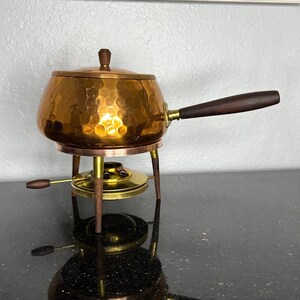 May include: A copper fondue pot with a hammered finish and a wooden handle sits on a brass stand with three legs. The stand has a small burner for melting chocolate or cheese.