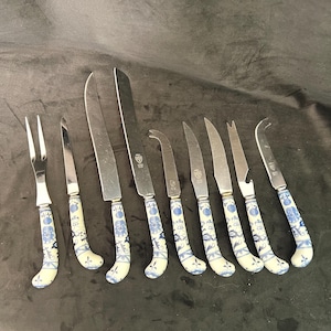 May include: A collection of cheese knives and a fork with stainless steel blades and white handles decorated with blue floral patterns. The set includes various knife shapes for different cheeses.
