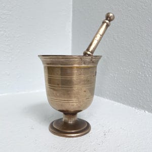 May include: A vintage brass mortar and pestle set. The mortar has a wide, cylindrical bowl with horizontal ridges and a pedestal base. The pestle is cylindrical with a rounded handle. The set is made of a warm, golden-toned brass.