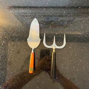 May include: Two kitchen utensils, a silver cake server with a black and orange handle and a silver three-pronged fork with a black handle.