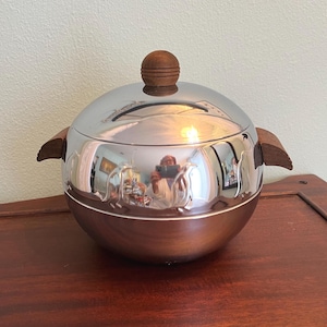 May include: A silver-colored ice bucket with a wooden lid and handles. The round bucket has a shiny, reflective surface and a dark brown wooden base. The lid is topped with a small, round wooden knob.