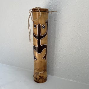 May include: A brown bamboo tube with a black and white design of a stylized figure with two eyes. The tube has a string tied around the top.