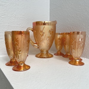 May include: An iridescent orange glass pitcher and matching glasses. The pitcher has a handle and a wide base. The glasses have a tulip shape and are decorated with a floral pattern. The set is displayed on a white surface.