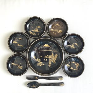 May include: A black and gold Japanese-style serving set featuring a large plate, several small bowls, a fork, and a spoon. The set has a gold mountain and landscape design.