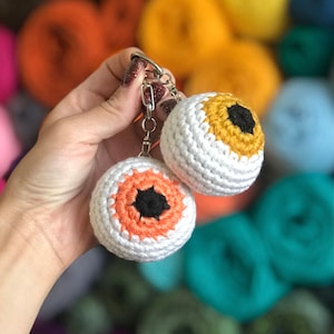 May include: Two crocheted eyeball keyrings with black pupils. One eyeball is orange and white, the other is yellow and white.