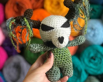 May include: A handmade crochet doll with a skull-like head, black eyes, and a heart-shaped nose. The doll has brown antlers, green and orange accents, and is holding a small orange pumpkin. The doll's body is green.