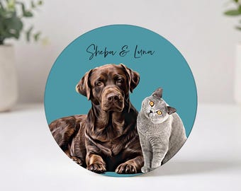 May include: A circular plaque featuring a digital illustration of a brown Labrador dog and a grey cat against a teal background. The text Sheba & Luna is printed in black script above the animals. The plaque has a smooth, flat surface.