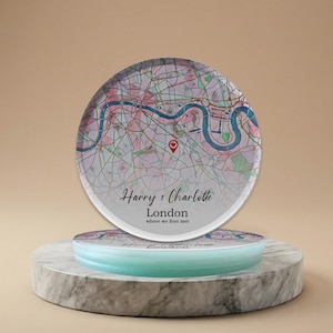Puede incluir: Un mapa redondo de cristal de Londres, con un marcador de corazón rojo. El mapa está impreso con un efecto acuarela en tonos rosas, morados y azules. El texto "Harry & Charlotte, London, where we first met" está impreso.