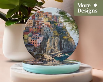 May include: A round coaster featuring a colorful coastal village scene. The coaster is displayed on a marble and light blue stand, with a potted plant in the background. The text "More Designs" is visible.