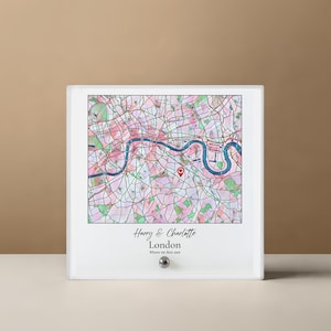 May include: A framed map of London, featuring a watercolor-style design in pink, green, and blue. The River Thames is prominently displayed. The text "Harry & Charlotte London Where we first met" is at the bottom. A small, silver knob is at the bottom.