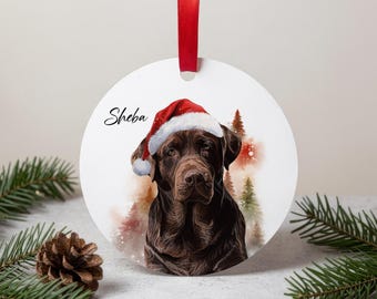 May include: A circular Christmas decoration with a brown dog wearing a Santa hat. The dog's name, "Sheba," is written in script. A red ribbon is attached for hanging. The background features a watercolour forest scene.
