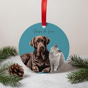 Può includere: Ornamento rotondo color teal con un ritratto di un cane marrone e un gatto grigio. Il cane è un Labrador Retriever marrone e il gatto è un British Shorthair grigio. L'ornamento ha un nastro rosso e il testo "Sheba & Luna".