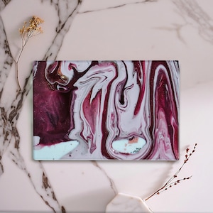 May include: Rectangular decorative art piece with a swirling pattern of burgundy, white, and gold. The abstract design resembles flowing paint or marble. The artwork is displayed on a marble surface.