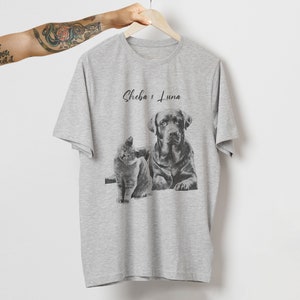 May include: A heather gray t-shirt featuring a black and white illustration of a cat and a dog. The cat is sitting, and the dog is lying down. The text "Sheba & Luna" is above the illustration.