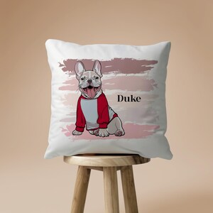 May include: White square throw pillow featuring a cartoon illustration of a French Bulldog wearing a red and white hoodie. The dog is sitting and smiling with its tongue out. The name "Duke" is printed in black. Pink and mauve brushstrokes are in the background.