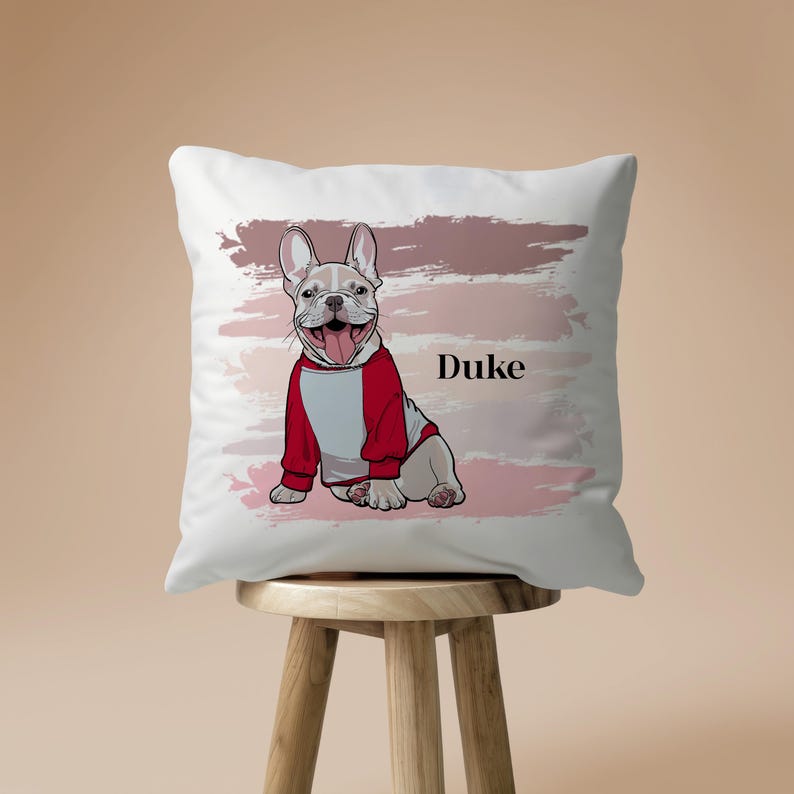 May include: White square throw pillow featuring a cartoon illustration of a French Bulldog wearing a red and white hoodie. The dog is sitting and smiling with its tongue out. The name "Duke" is printed in black. Pink and mauve brushstrokes are in the background.