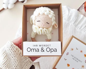 Puede incluir: Un sonajero de oveja de ganchillo con un anillo de madera en una caja de regalo de cartón. El inserto de la caja tiene el texto "IHR WERDET Oma & Opa" y la tapa muestra una corona floral con el texto "EINE KLEINIGKEIT FÜR DICH" en negro.
