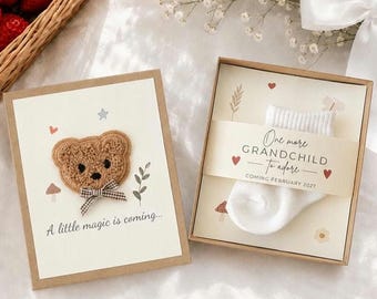 May include: A rectangular card featuring a brown fuzzy bear patch with a gingham bow and the text A little magic is coming, alongside a gift box containing a white baby sock and a band reading One more GRANDCHILD To adore.
