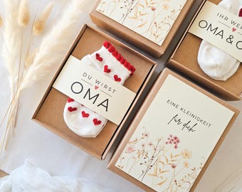 Puede incluir: Tres cajas de regalo de cartón marrón que contienen calcetines de bebé blancos con patrones de corazones rojos. Cada caja presenta una banda de papel con texto en alemán que incluye 'DU WIRST OMA' y 'EINE KLEINIGKEIT für dich', junto a ilustraciones florales.