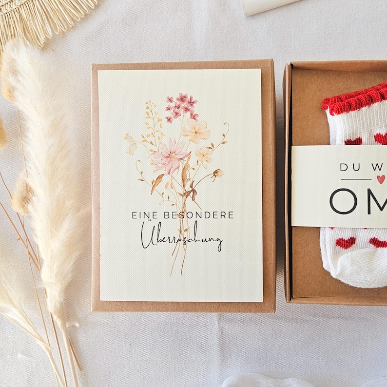 Puede incluir: Una caja de regalo de cart&oacute;n marr&oacute;n que contiene una tarjeta blanca con una ilustraci&oacute;n floral en acuarela y el texto EINE BESONDERE &Uuml;berraschung, junto a un par de calcetines blancos con corazones rojos y una banda que dice DU W OMA.