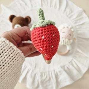 Puede incluir: Un sonajero de fresa tejido a ganchillo con un cuerpo rojo brillante, semillas bordadas en blanco y un tallo verde texturizado. La fresa est&aacute; unida a un anillo de madera lisa de color claro, dise&ntilde;ado como juguete de mano para beb&eacute;s y ni&ntilde;os.