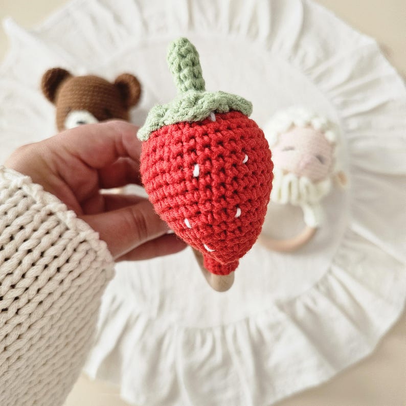 Puede incluir: Un sonajero de fresa tejido a ganchillo con un cuerpo rojo brillante, semillas bordadas en blanco y un tallo verde texturizado. La fresa est&aacute; unida a un anillo de madera lisa de color claro, dise&ntilde;ado como juguete de mano para beb&eacute;s y ni&ntilde;os.