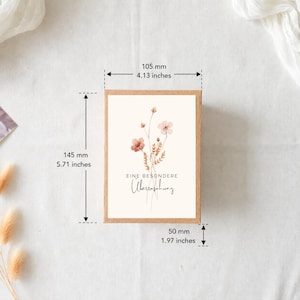 Puede incluir: Una caja de cart&oacute;n rectangular con una etiqueta blanca que presenta una ilustraci&oacute;n floral en acuarela y el texto EINE BESONDERE &Uuml;berraschung. La caja mide 105 mm de ancho, 145 mm de alto y 50 mm de profundidad seg&uacute;n las medidas.