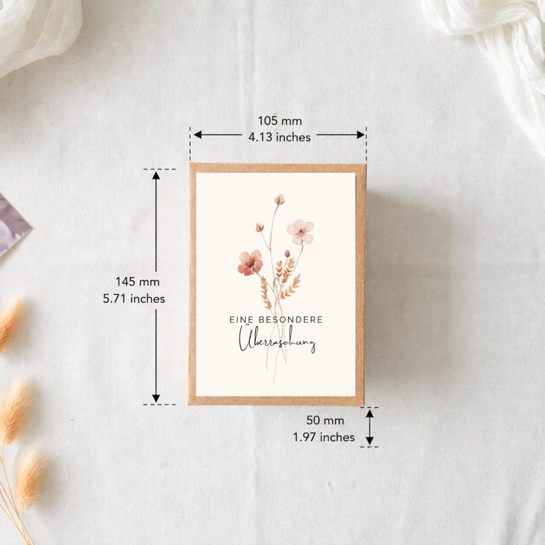 Puede incluir: Una caja de cart&oacute;n rectangular con una etiqueta blanca que presenta una ilustraci&oacute;n floral en acuarela y el texto EINE BESONDERE &Uuml;berraschung. La caja mide 105 mm de ancho, 145 mm de alto y 50 mm de profundidad seg&uacute;n las medidas.