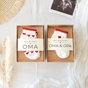 Puede incluir: Dos cajas de regalo de cart&oacute;n marr&oacute;n que contienen cada una un par de calcetines blancos con peque&ntilde;os corazones rojos y un borde con volantes rojos. La caja izquierda tiene una banda con DU WIRST OMA y la derecha con IHR WERDET OMA & OPA.