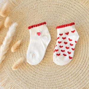 Puede incluir: Un par de calcetines tobilleros blancos con un borde rojo con volantes en el pu&ntilde;o. Un calcet&iacute;n presenta un peque&ntilde;o coraz&oacute;n rojo, mientras que el otro est&aacute; cubierto con un patr&oacute;n repetitivo de corazones rojos peque&ntilde;os. Tejido de punto acanalado.