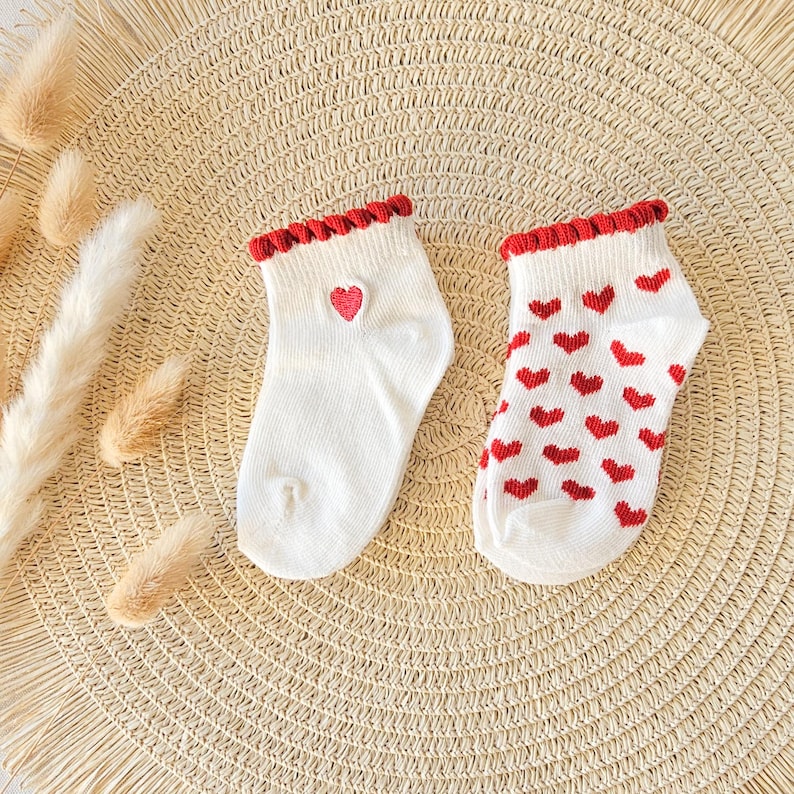 Puede incluir: Un par de calcetines tobilleros blancos con un borde rojo con volantes en el pu&ntilde;o. Un calcet&iacute;n presenta un peque&ntilde;o coraz&oacute;n rojo, mientras que el otro est&aacute; cubierto con un patr&oacute;n repetitivo de corazones rojos peque&ntilde;os. Tejido de punto acanalado.