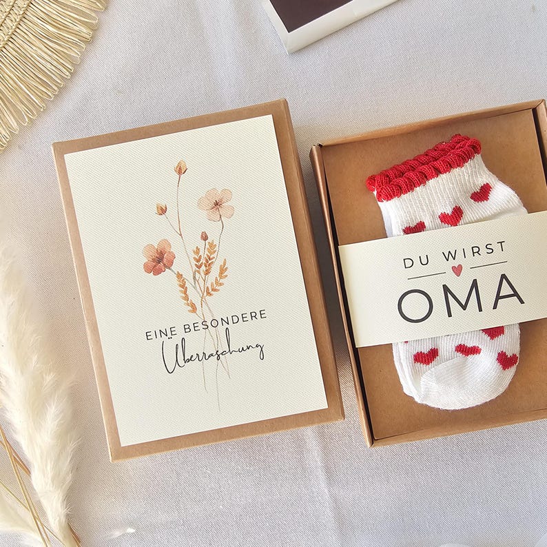 Puede incluir: Una caja de regalo de cart&oacute;n marr&oacute;n que contiene un calcet&iacute;n blanco con corazones rojos y un pu&ntilde;o acanalado rojo, envuelto en una banda de papel con el texto DU WIRST OMA. Junto a ella, una tarjeta con una ilustraci&oacute;n floral y el texto EINE BESONDERE &Uuml;berraschung.