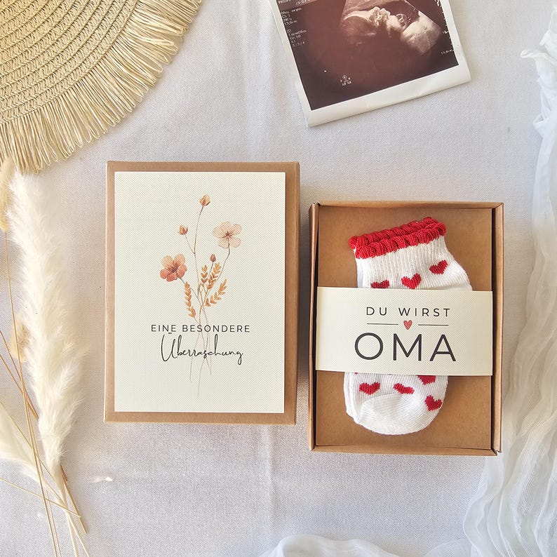 Puede incluir: Una caja de regalo de cart&oacute;n marr&oacute;n que contiene calcetines de beb&eacute; blancos con peque&ntilde;os corazones rojos y una banda de papel con el texto DU WIRST OMA y un coraz&oacute;n. Al lado, una tarjeta con una ilustraci&oacute;n floral y el texto EINE BESONDERE &Uuml;berraschung.