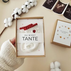 Puede incluir: Un par de calcetines de beb&eacute; color crema con borde rojo y un peque&ntilde;o detalle de coraz&oacute;n rojo, presentados en una caja de regalo de cart&oacute;n. Una banda de papel blanco sobre los calcetines presenta el texto negro DU WIRST TANTE.