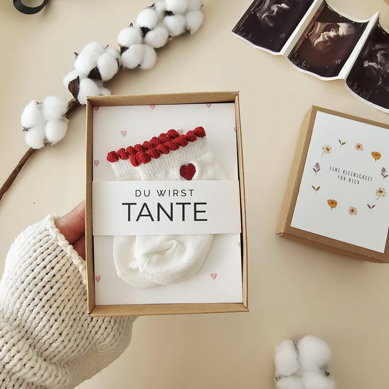 Puede incluir: Un par de calcetines de beb&eacute; color crema con borde rojo y un peque&ntilde;o detalle de coraz&oacute;n rojo, presentados en una caja de regalo de cart&oacute;n. Una banda de papel blanco sobre los calcetines presenta el texto negro DU WIRST TANTE.
