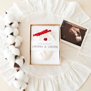 Puede incluir: Una caja de regalo de cart&oacute;n que contiene un par de calcetines de beb&eacute; blancos con borde rojo y un peque&ntilde;o coraz&oacute;n rojo, envueltos en una banda de papel con el texto IHR WERDET UROMA & UROPA, junto a una ecograf&iacute;a y una rama de algod&oacute;n.