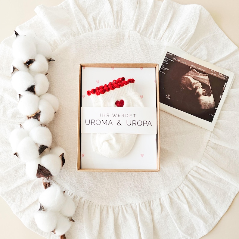 Puede incluir: Una caja de regalo de cart&oacute;n que contiene un par de calcetines de beb&eacute; blancos con borde rojo y un peque&ntilde;o coraz&oacute;n rojo, envueltos en una banda de papel con el texto IHR WERDET UROMA & UROPA, junto a una ecograf&iacute;a y una rama de algod&oacute;n.