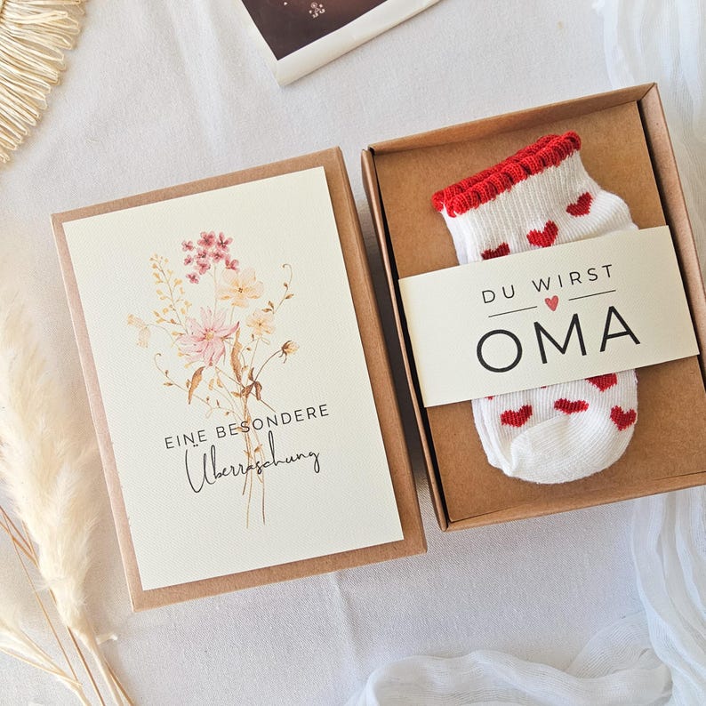 Puede incluir: Una caja de regalo de cart&oacute;n marr&oacute;n que contiene un calcet&iacute;n blanco con peque&ntilde;os corazones rojos y un borde acanalado rojo, envuelto en una banda de papel con el texto DU WIRST OMA. Junto a ella, una tarjeta con flores y el texto EINE BESONDERE &Uuml;berraschung.