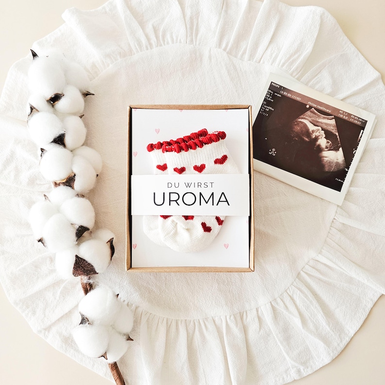 Puede incluir: Un par de calcetines de beb&eacute; blancos con peque&ntilde;os corazones rojos y un borde festoneado rojo, presentados en una caja de cart&oacute;n con una banda de papel que dice DU WIRST UROMA, junto a una ecograf&iacute;a y una rama de algod&oacute;n blanco.
