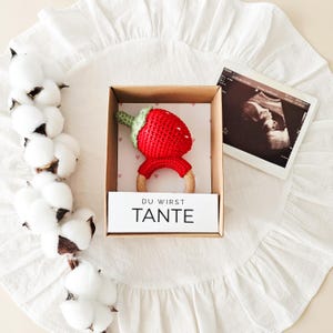 Puede incluir: Una caja de regalo de cart&oacute;n que contiene un sonajero de fresa de ganchillo rojo con un tallo verde unido a un anillo de madera. Una tarjeta blanca dentro de la caja presenta el texto alem&aacute;n DU WIRST TANTE en letras negras sobre corazones rosas.